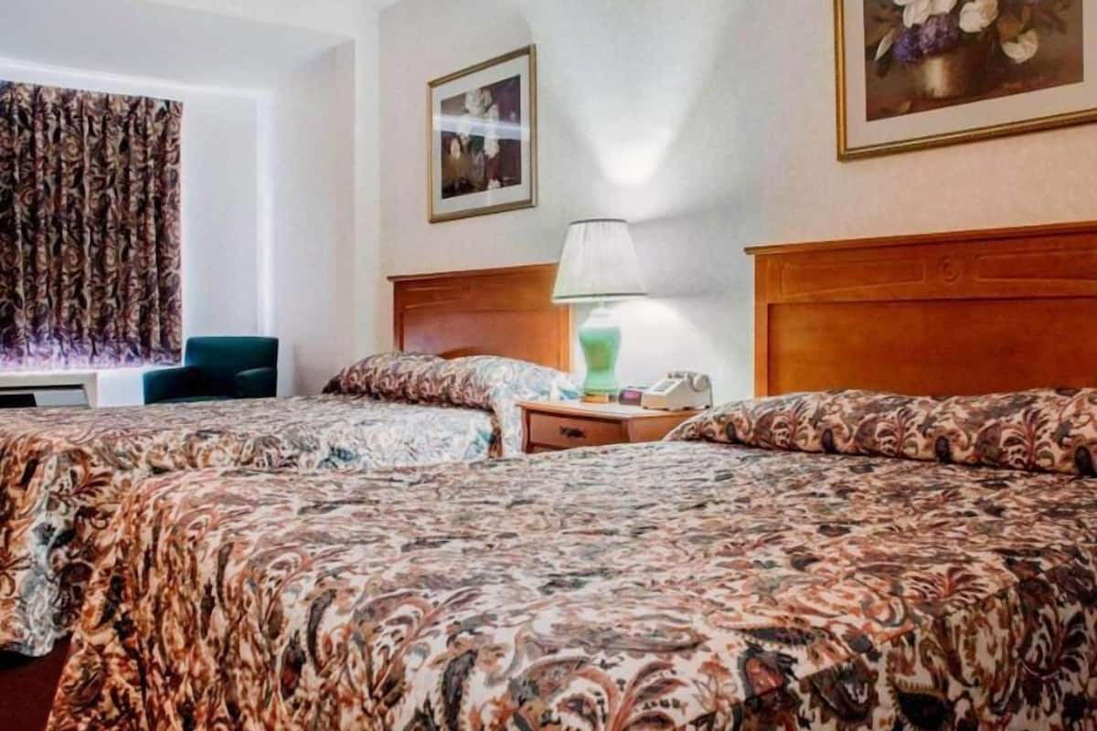 Habitación de hotel con dos camas, una mesita de noche con lámpara, una silla y un ventanal con cortinas.