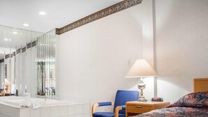 A hotel room with a bed, a chair, a nightstand with a lamp, a bathroom with a bathtub, and a large mirror.