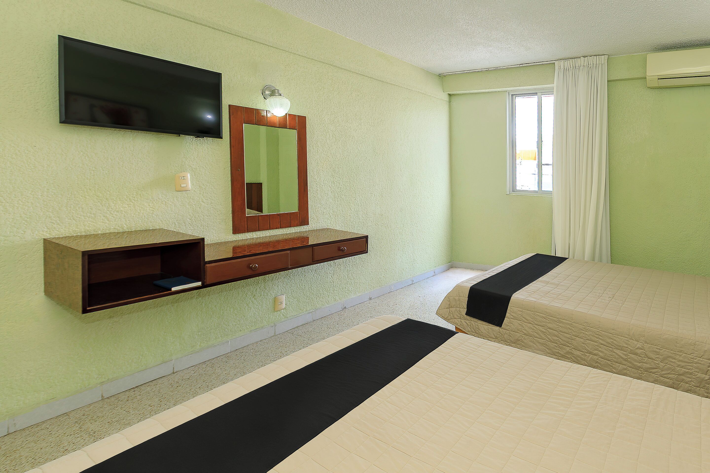 Quarto de hotel com cama, televisão fixada na parede, mesa de cabeceira e janela com cortinas.