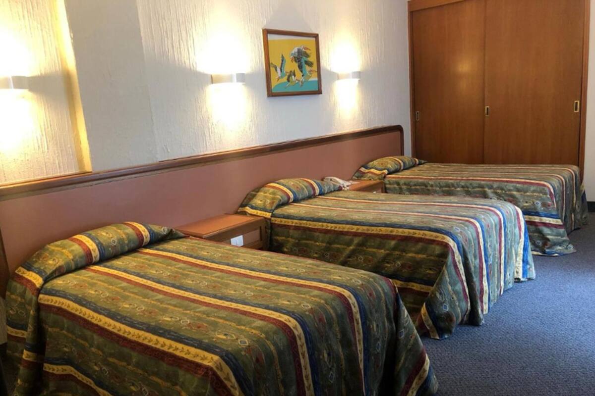 A hotel room with four single beds, each with patterned bedding, a wooden headboard, and a wall-mounted light fixture.