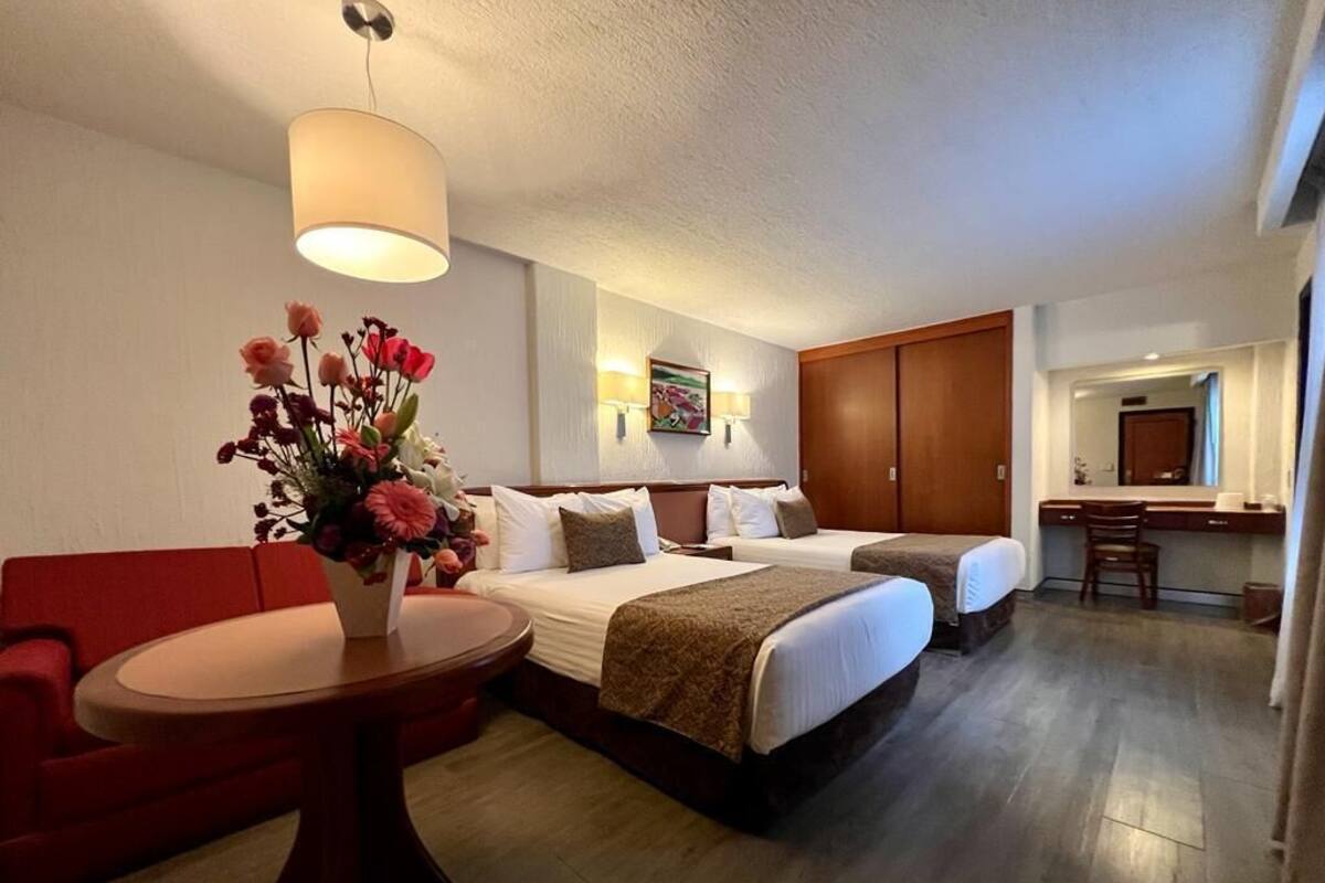 A hotel room with two beds, a desk, a chair, a red sofa, and a table with a vase of flowers.