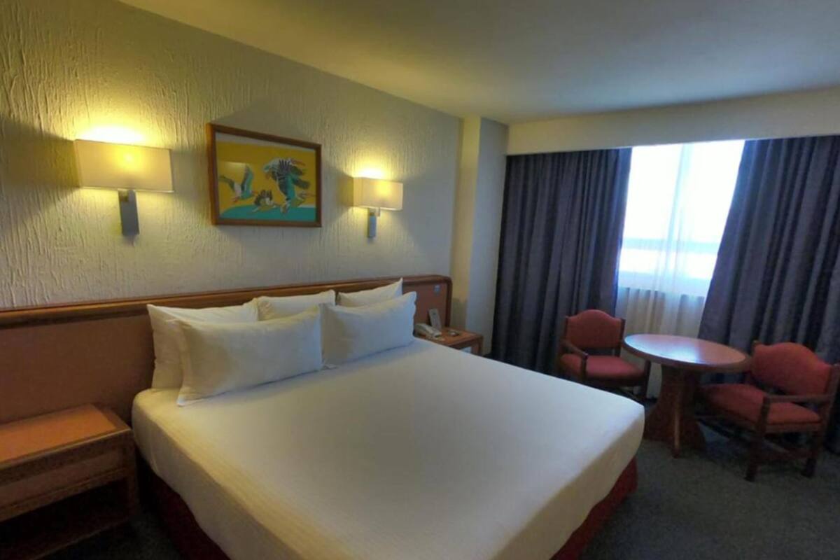 A hotel room with a large bed, two bedside lamps, a painting on the wall, a small table with two chairs, and a window with curtains.