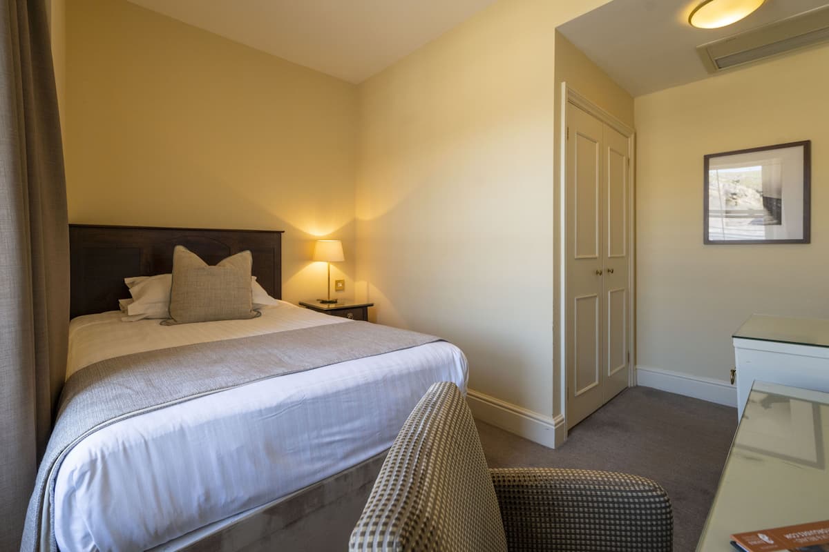 A hotel room with a neatly made bed, a bedside table with a lamp, a chair, and a desk with a chair.