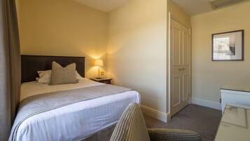 A hotel room with a neatly made bed, a bedside table with a lamp, a chair, and a desk with a chair.