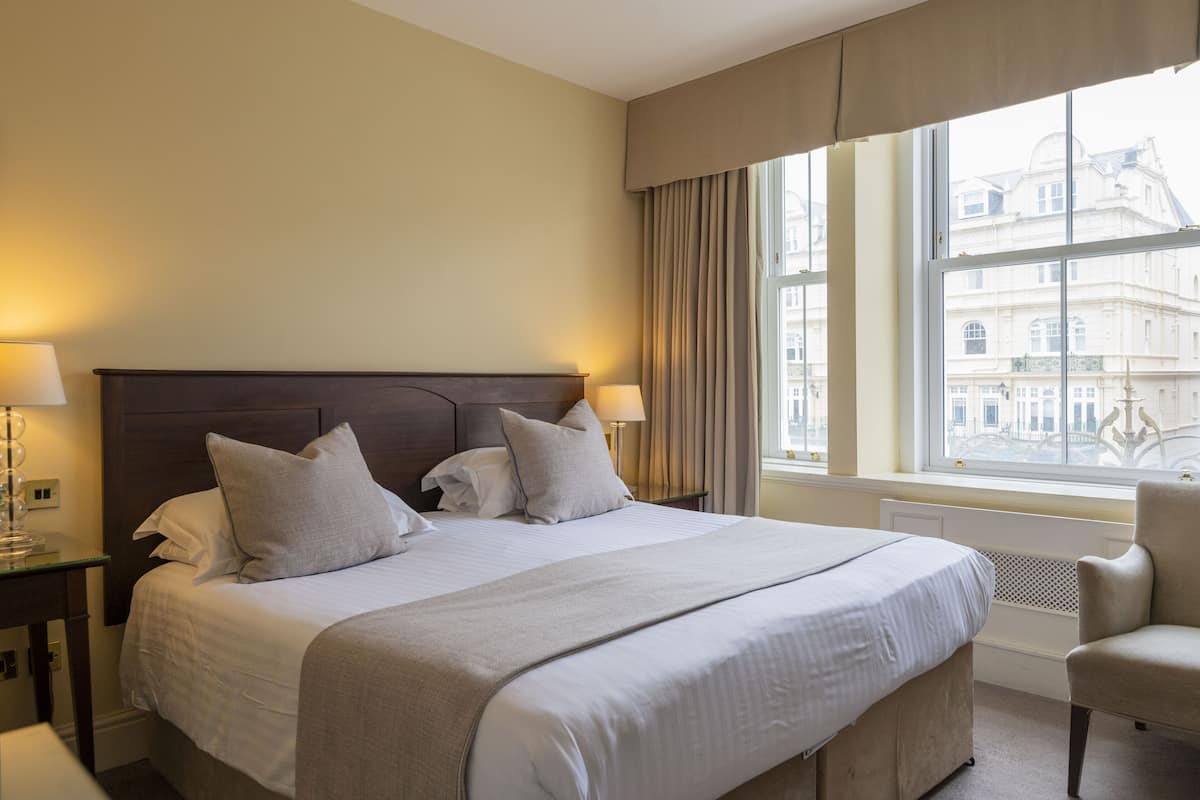 A hotel room with a large bed, two bedside lamps, a chair, and a window with a city view.
