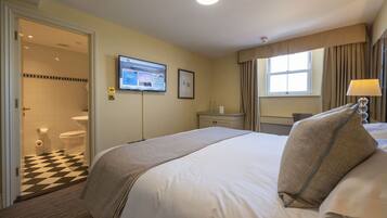 A hotel room with a bed, a desk, a chair, a window with curtains, a television on the wall, and a bathroom with a checkered floor.