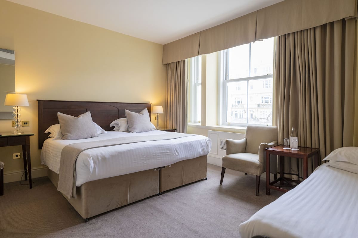 A hotel room with a large bed, two bedside tables with lamps, a chair, and a view of buildings through the window.