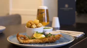 A plate of fried fish with lemon and greens, accompanied by a bowl of chips and a small bowl of sauce, set on a table with a menu and a glass of beer in the background.
