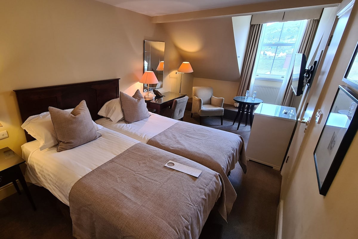 A hotel room with two beds, a desk, a chair, and a small table with a lamp.