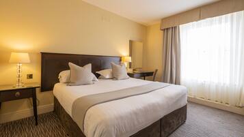 A hotel room with a large bed, two bedside tables with lamps, a nightstand, a mirror, and a window with curtains.