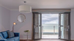 A modern room with a blue sofa, a small table with a plant, a round mirror, and a large glass door leading to a balcony with a view of the sea. - St George's Hotel (Llandudno)