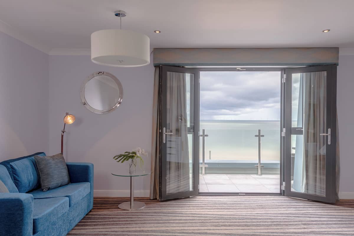 A modern room with a blue sofa, a small table with a plant, a round mirror, and a large glass door leading to a balcony with a view of the sea.