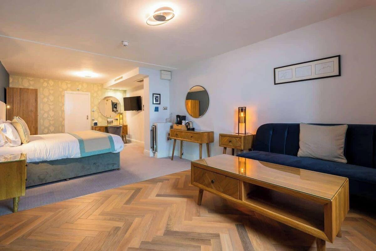 A hotel room with a bed, a sofa, a wooden coffee table, a desk, a mirror, and a television.
