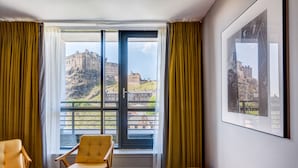 A hotel room with a large window offering a view of a historic building and a balcony. There are two chairs and a bench in the room.