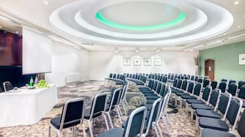 Un salón de conferencias con filas de sillas, una mesa blanca con botellas de agua y una pantalla para proyector.
