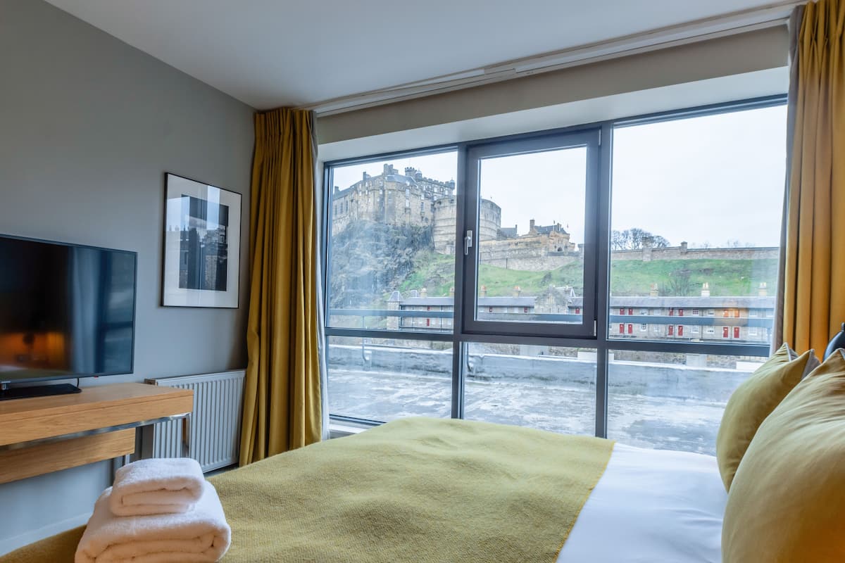 Habitación de hotel con cama, televisor, un cuadro enmarcado y ventana con vista a un castillo.