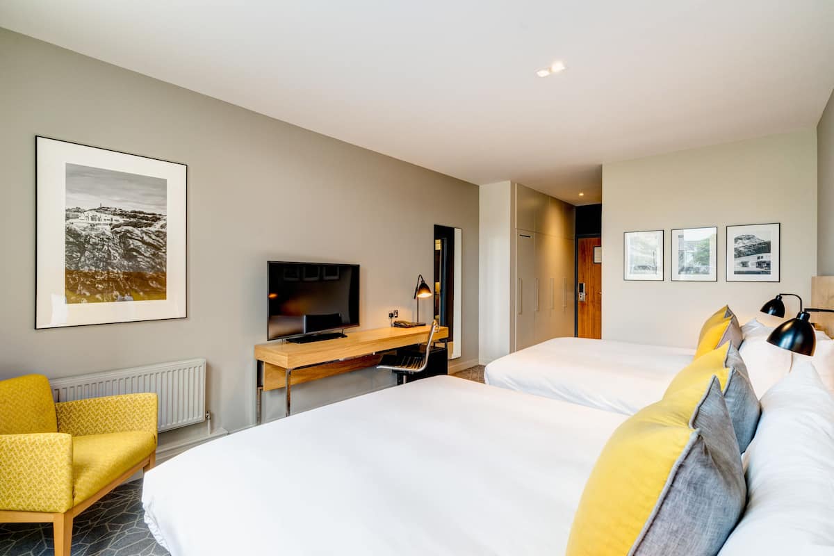Habitación de hotel con dos camas, un escritorio con televisor, una silla y cuadros en la pared.