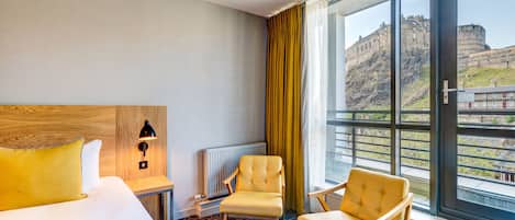Une chambre d’hôtel avec un grand lit, deux fauteuils, une table de chevet, une lampe et une vue sur un bâtiment historique.