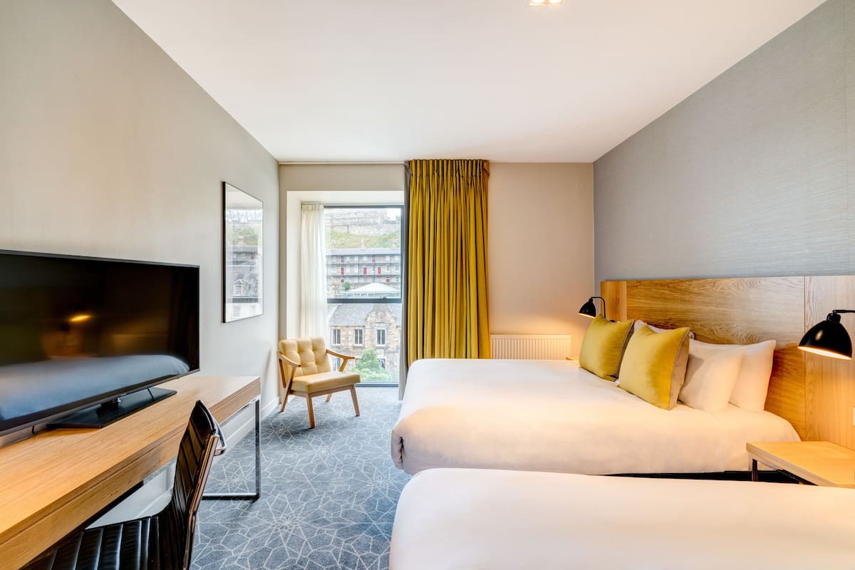 Habitación de hotel con una cama grande, un escritorio con televisor, una silla y vistas a un paisaje urbano a través de una ventana con cortinas amarillas.