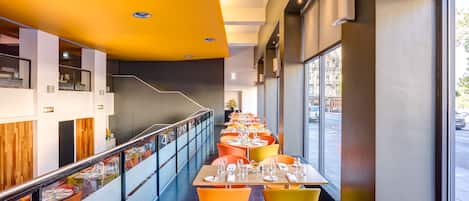 A modern dining area with long tables, orange chairs, and a view of the street through large windows.