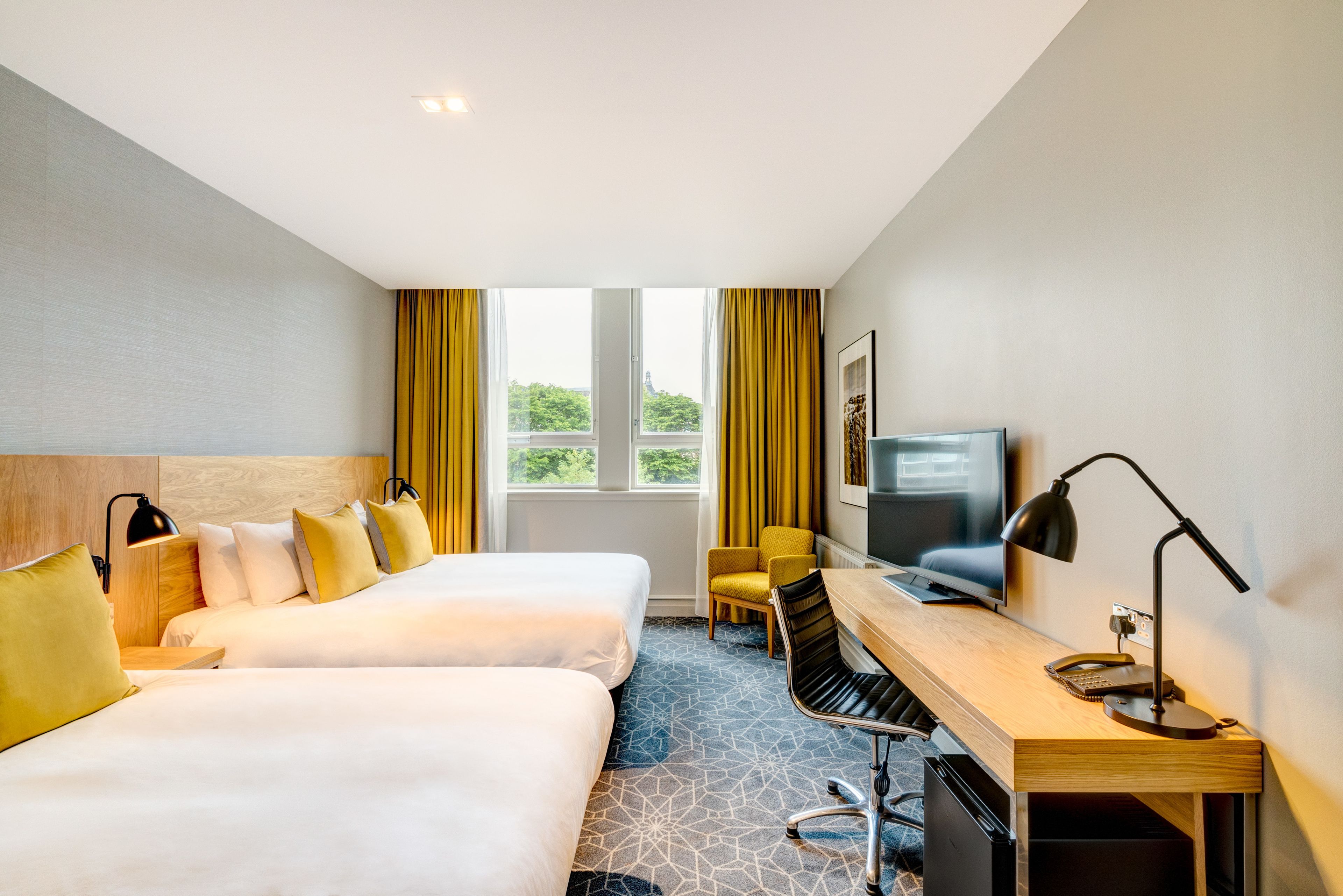 Ein Hotelzimmer mit zwei Betten, einem Schreibtisch, einem Stuhl, einem Fernseher und einem Fenster mit Vorhängen.