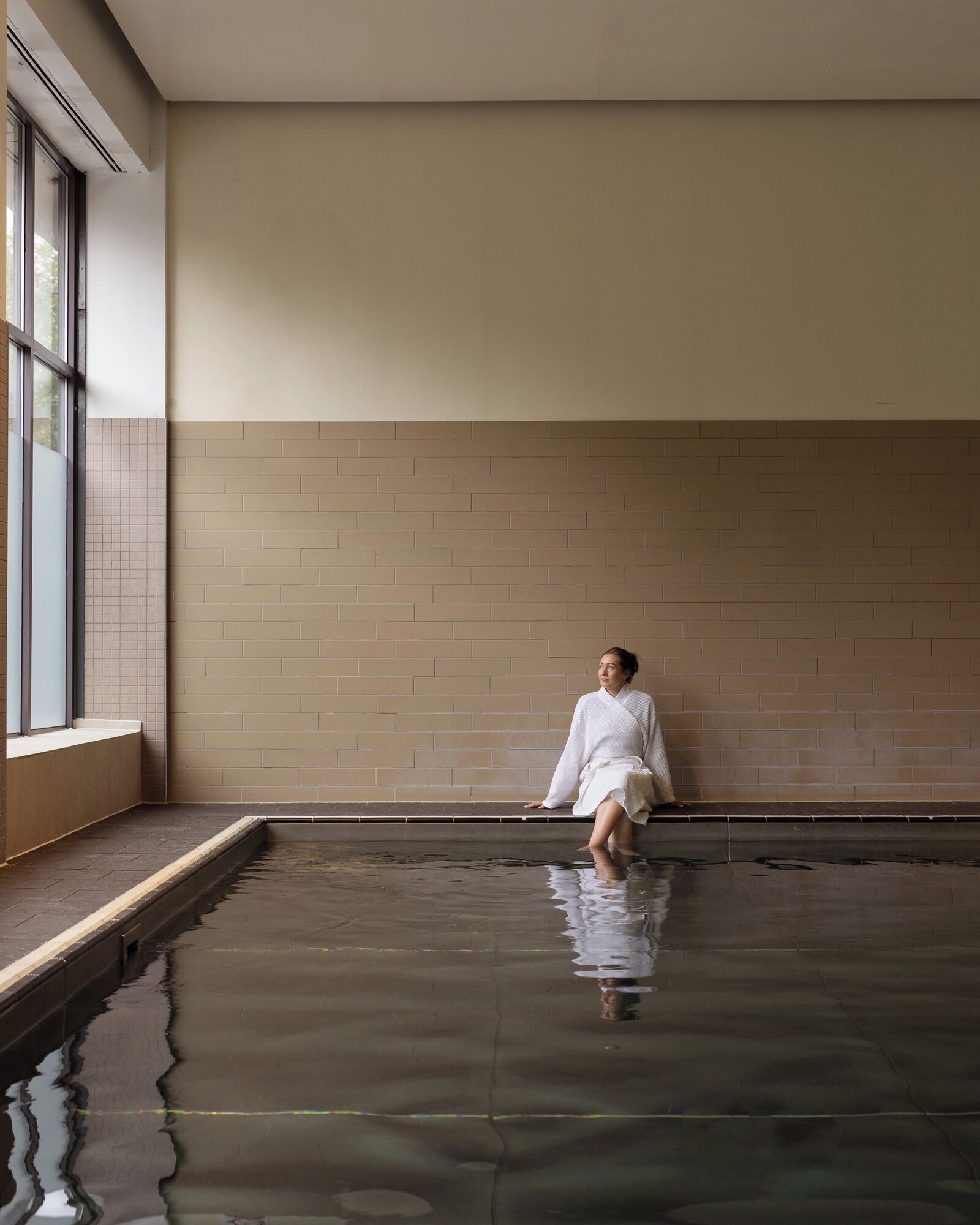 A person sitting by a pool in a room with a tiled wall and a window.