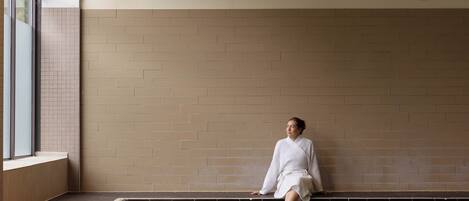 A person sitting by a pool in a room with a tiled wall and a window.