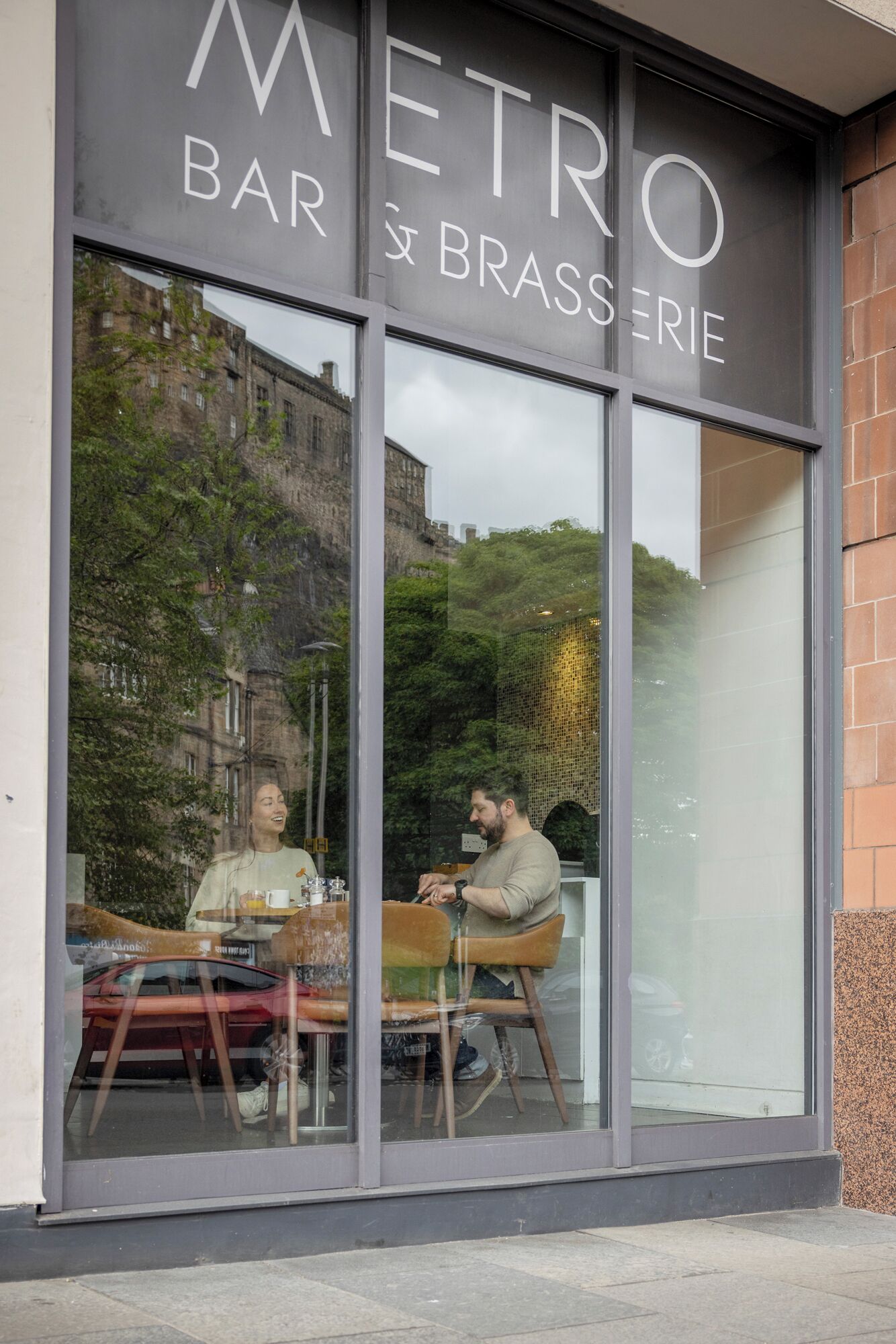 Un restaurante con amplios ventanales, un letrero que dice 'METRO BAR & BRASSERIE', y reflejos de personas en el interior.