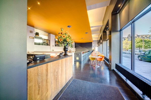 A modern hotel lobby with a reception desk, a large window offering a view of the street, and a seating area with tables and chairs. - Apex Grassmarket Hotel (Edinburgh)