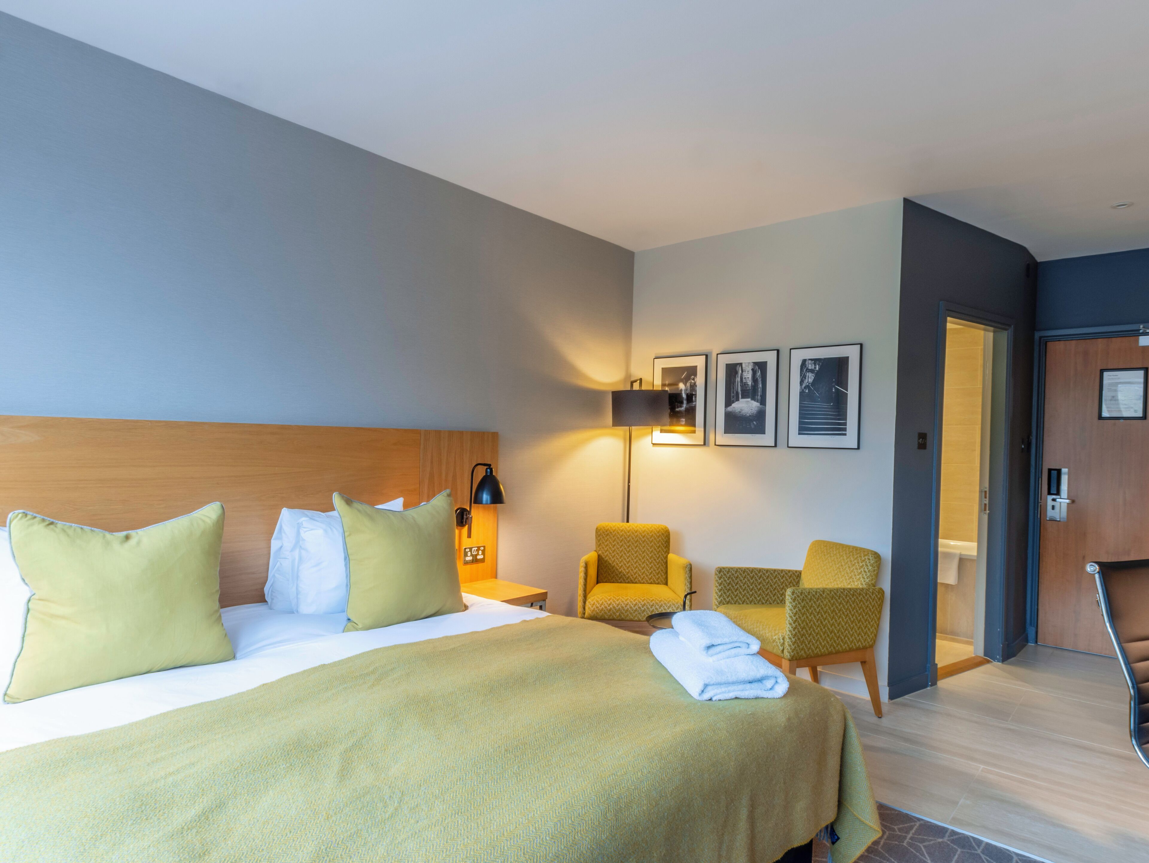 A hotel room with a bed, two chairs, a desk, a lamp, and framed pictures on the wall.
