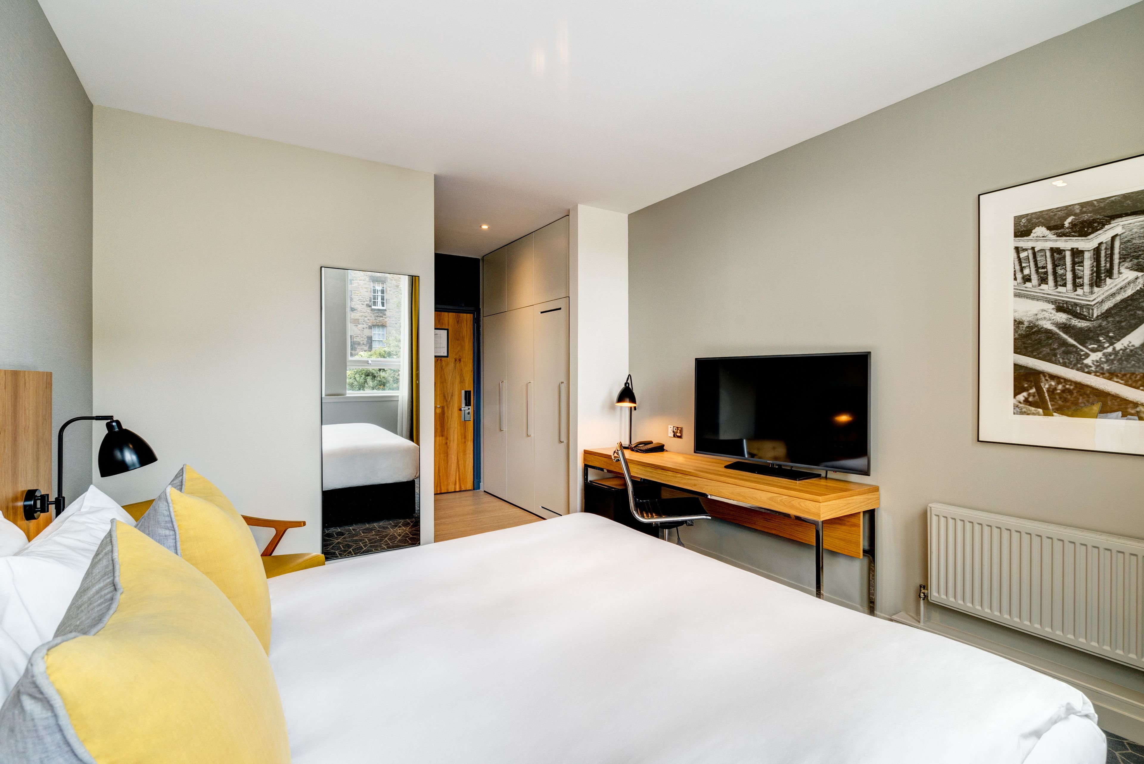Une chambre d’hôtel moderne avec un grand lit, un bureau équipé d’un ordinateur, une télévision et une fenêtre donnant sur un immeuble.