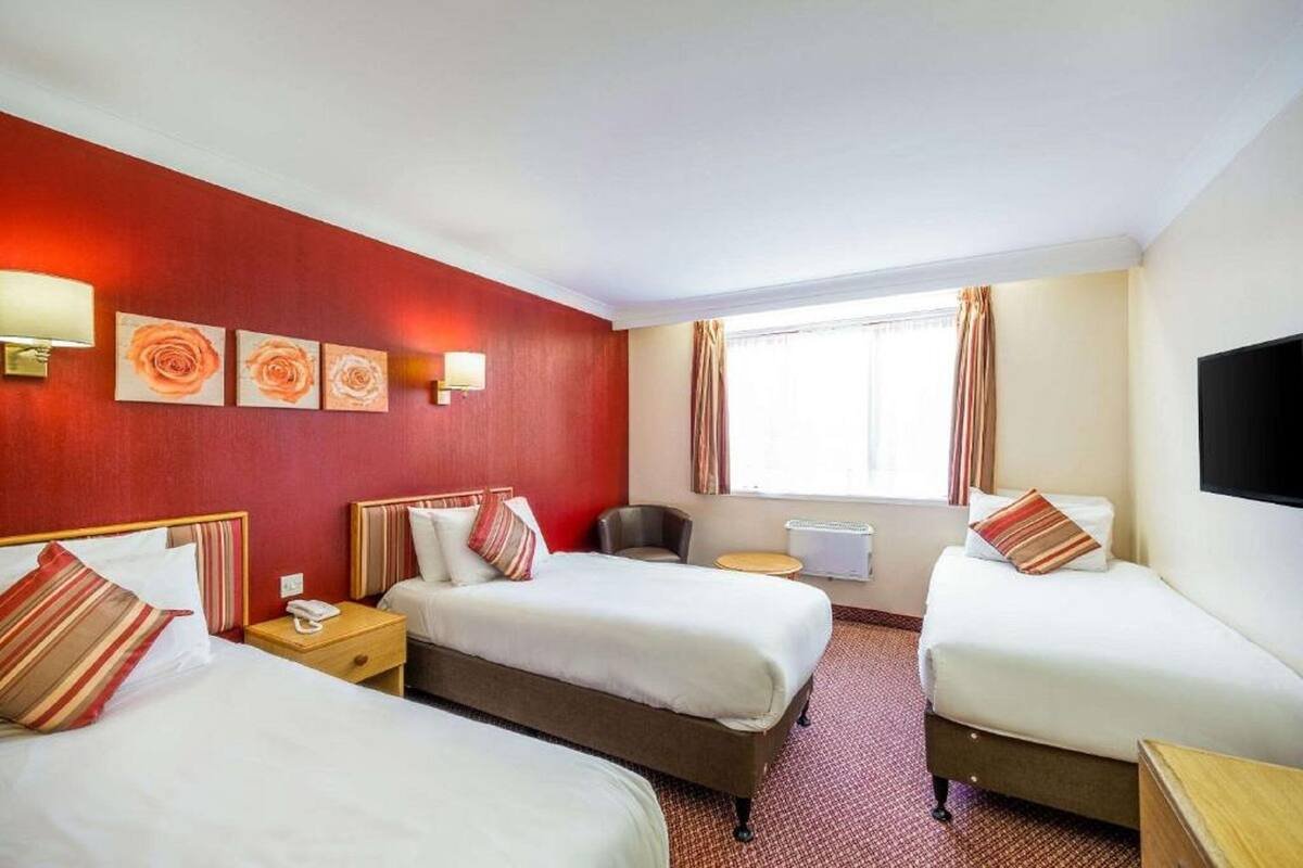 Ein Hotelzimmer mit drei Betten, einem Schreibtisch, einem Stuhl, einem Fernseher und Bildern an der Wand.