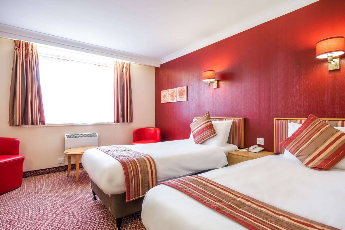 Ein Hotelzimmer mit zwei Betten, einem roten Sessel, einem kleinen Tisch und einem Fenster mit Vorhängen.