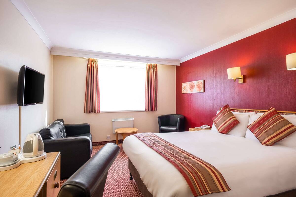 Ein Hotelzimmer mit Bett, Schreibtisch mit Wasserkocher, Stuhl, Fernseher und einem fensterbepflanzten Fenster.