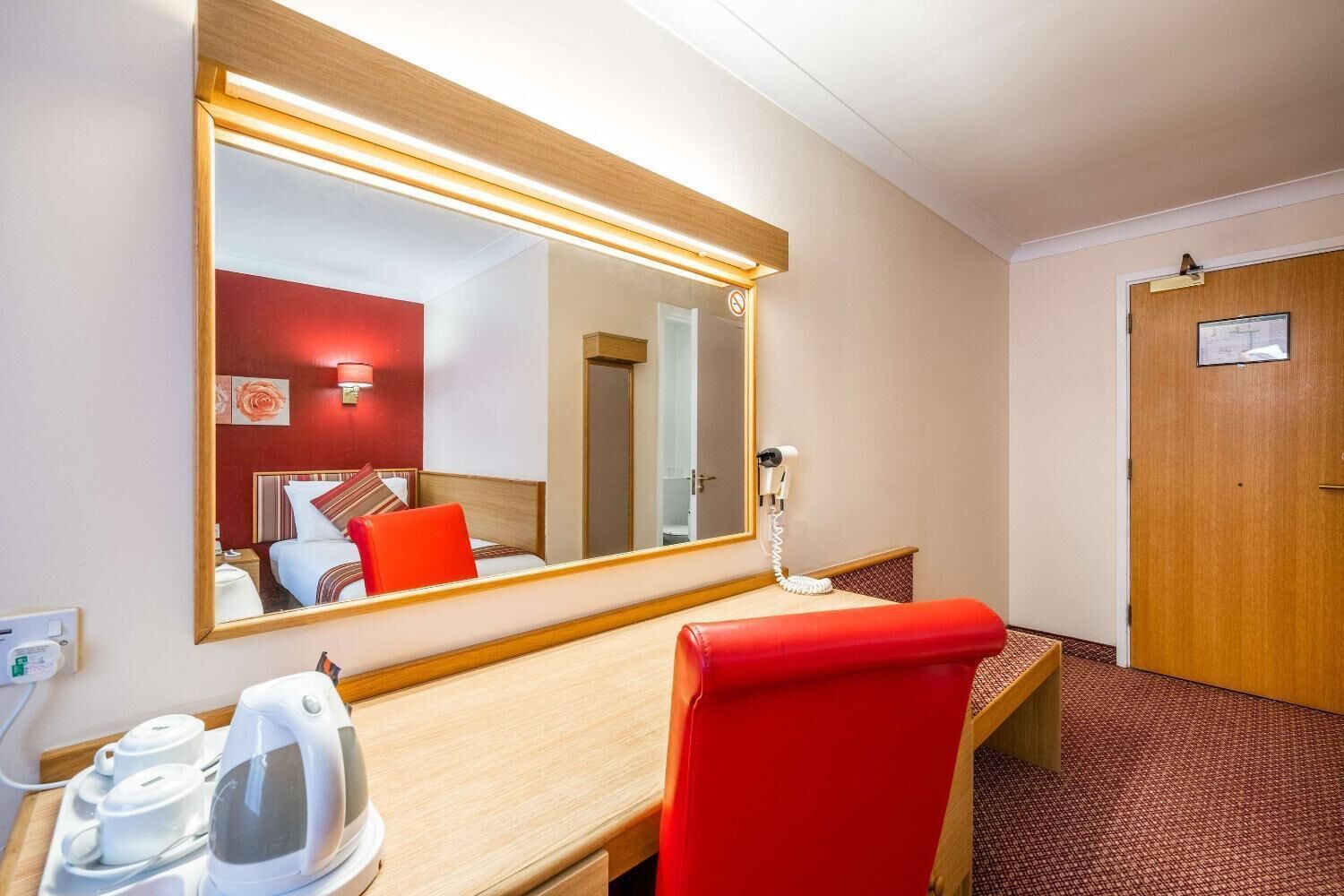 Habitación de hotel con un escritorio de madera, una silla roja, un espejo grande, un secador de pelo y una puerta con un cartel.