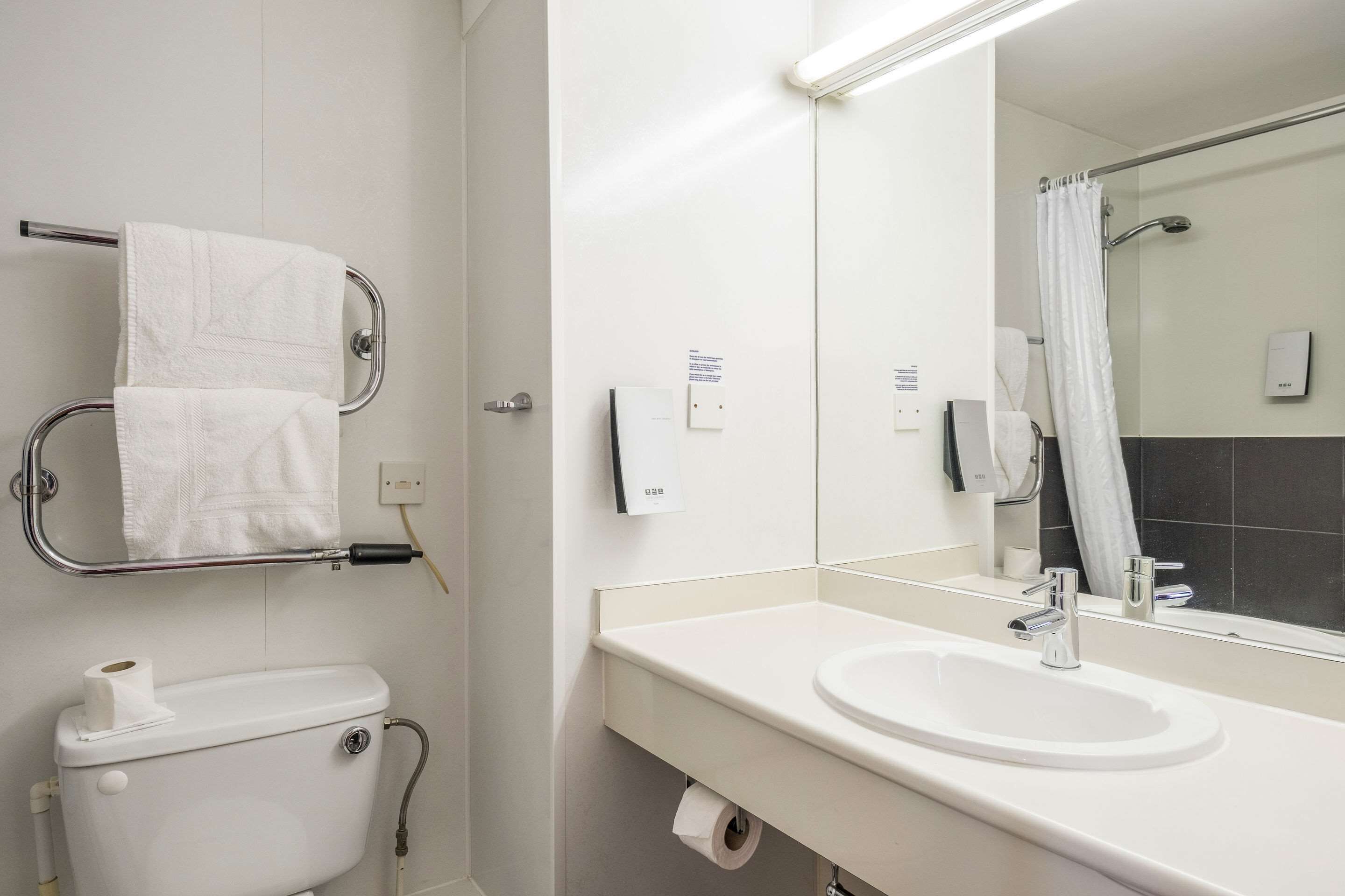 Un baño moderno con inodoro, lavamanos y espejo. Hay un perchero con dos toallas, un dispensador de toallas de papel y un dispensador de jabón.