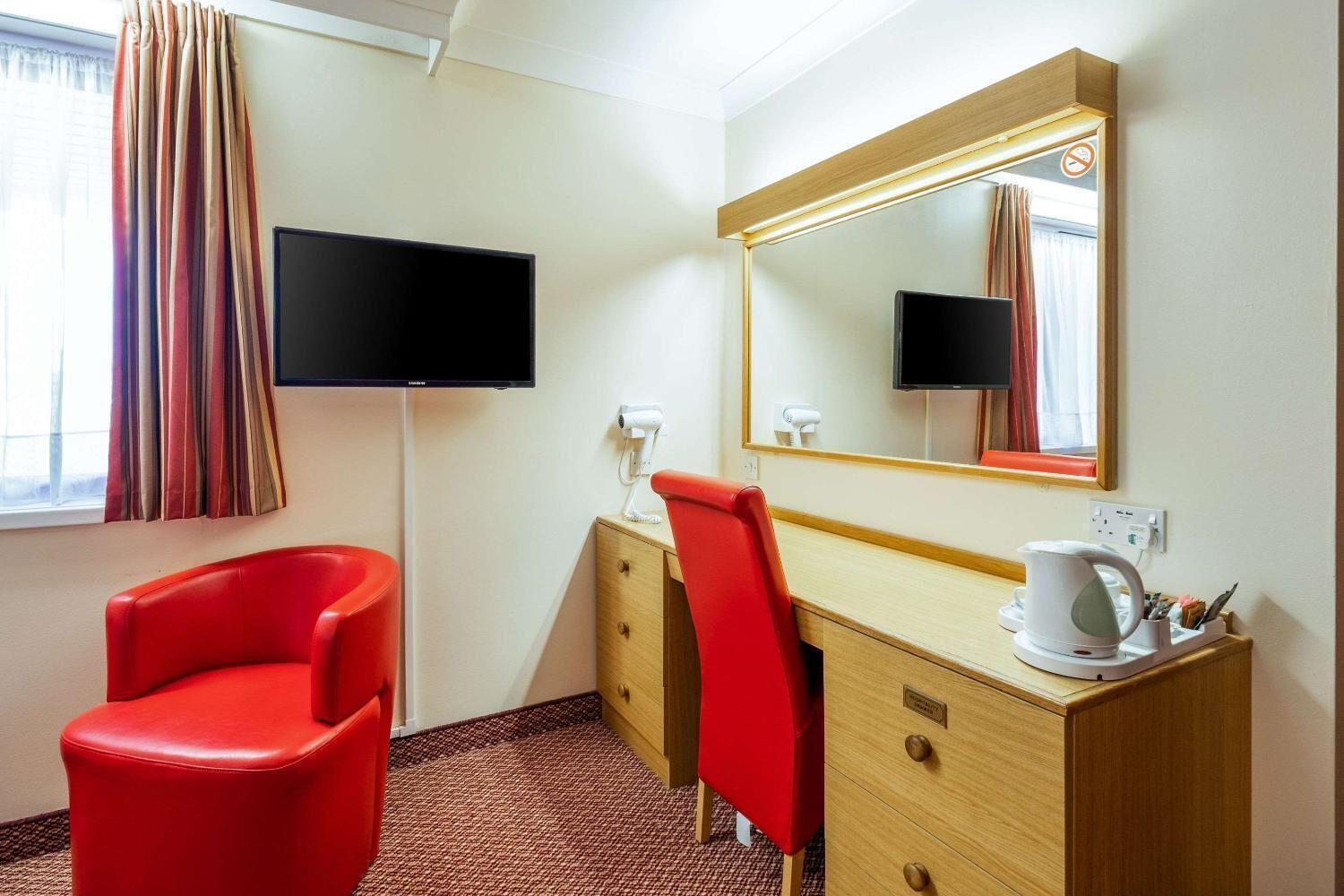A hotel room with a red chair, a wooden desk with a mirror, a flat-screen TV, and a coffee maker.