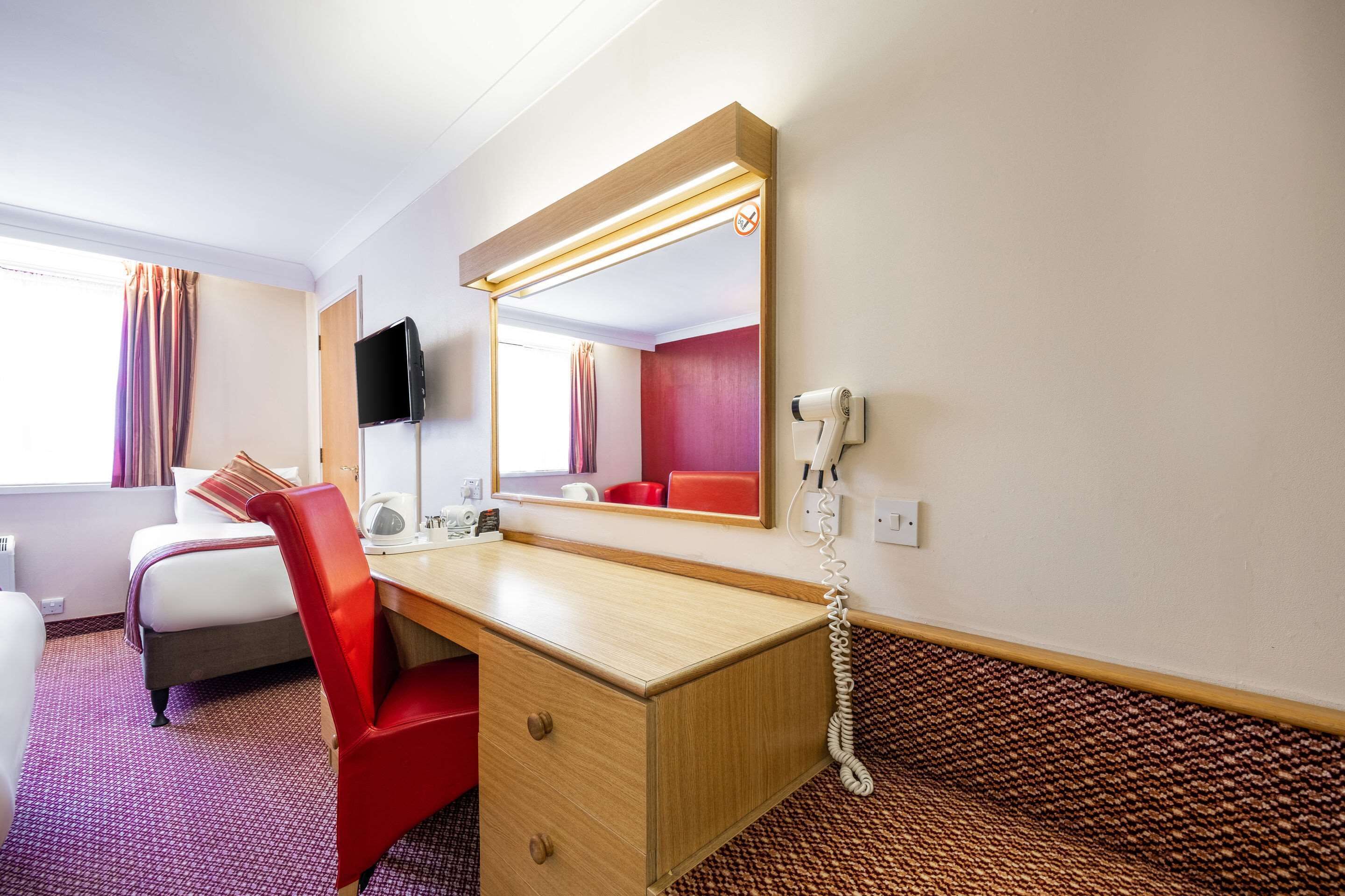 Ett hotellrum med ett skrivbord i trä, en röd stol, en säng, en tv och en hårtork.