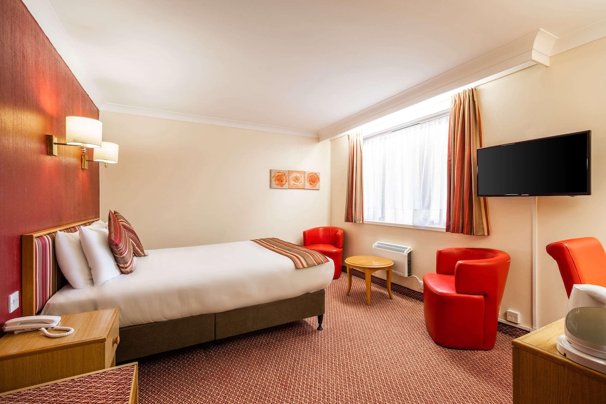 A hotel room with a bed, a TV, a red chair, a small table, and a wall-mounted lamp.
