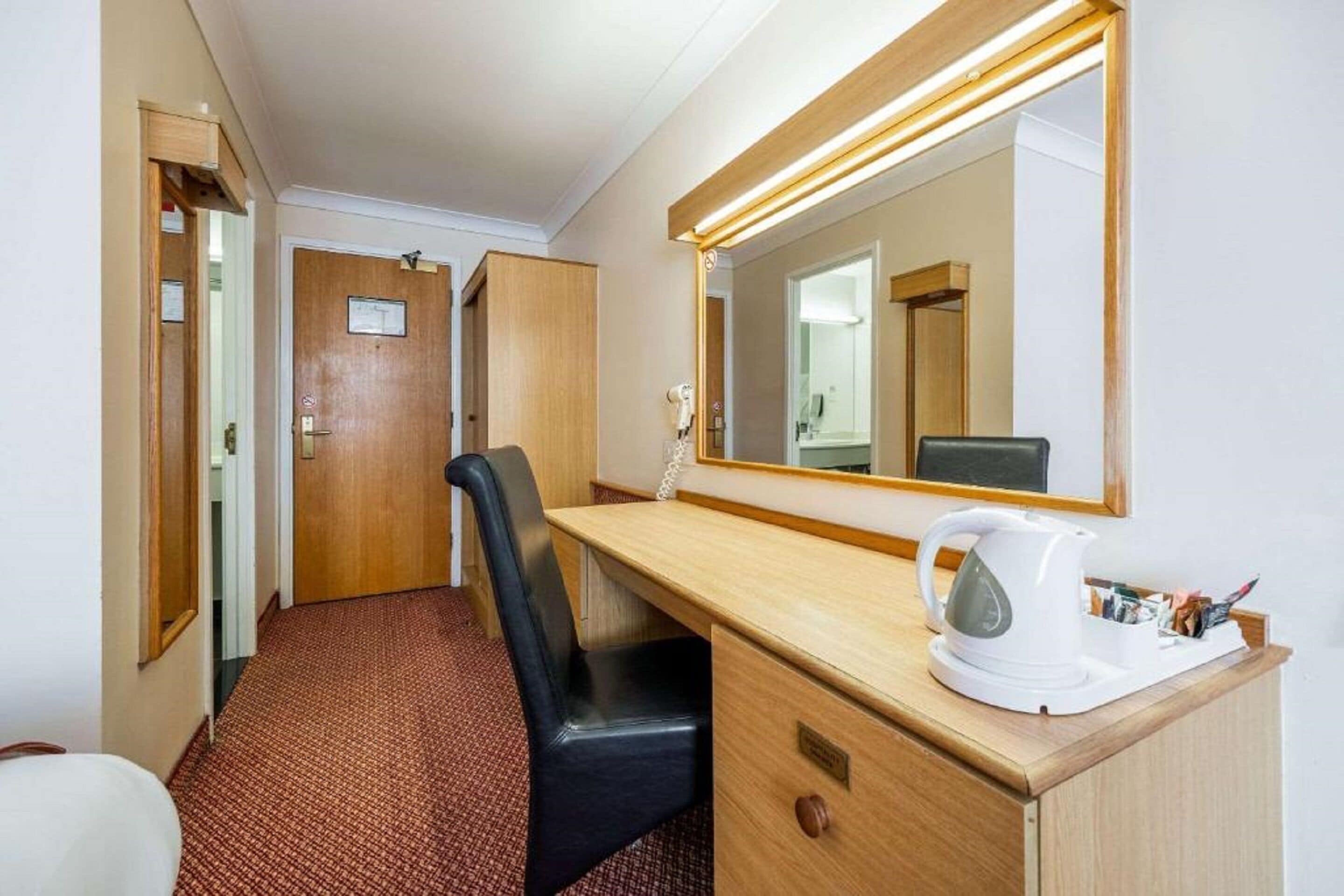 Une chambre d’hôtel avec un bureau en bois, une chaise de bureau noire, une bouilloire blanche et un grand miroir.