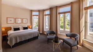 A hotel room with a large bed, two armchairs, a small table, and large windows with curtains. - The Resident Kensington (London)