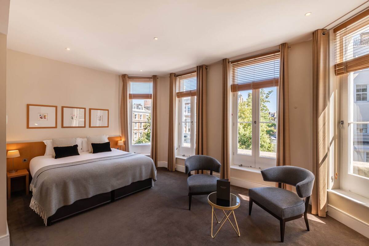 A hotel room with a large bed, two armchairs, a small table, and large windows with curtains.