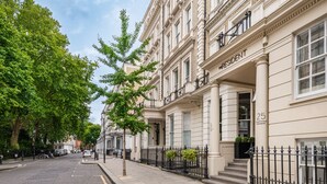 A row of traditional buildings with a classic architectural style, featuring a prominent entrance with a black iron fence and a sign indicating the address. - The Resident Kensington (London)