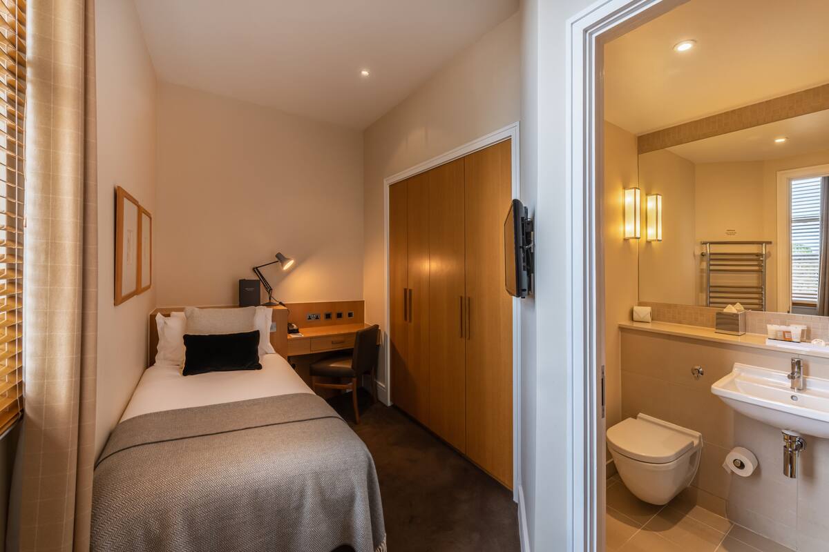 A compact hotel room with a bed, desk, chair, wardrobe, and a bathroom with a sink and toilet.