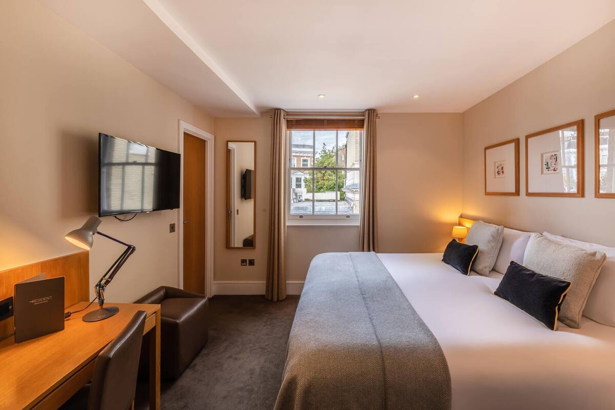 A hotel room with a large bed, a desk with a laptop, a TV mounted on the wall, and a window with curtains.