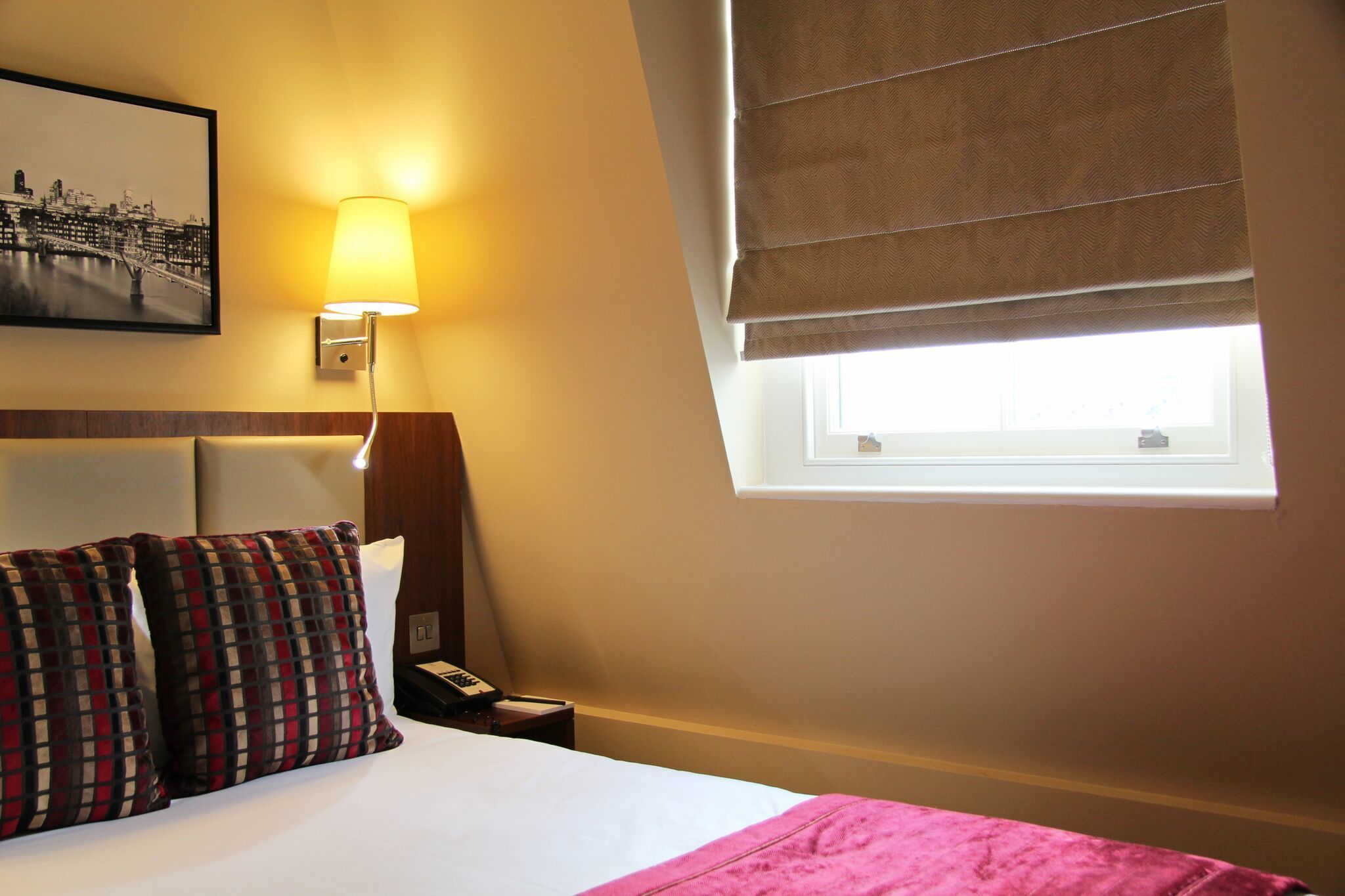 Une chambre d’hôtel avec un lit, une table de chevet, une lampe, une fenêtre avec un store et un tableau représentant un paysage urbain accroché au mur.