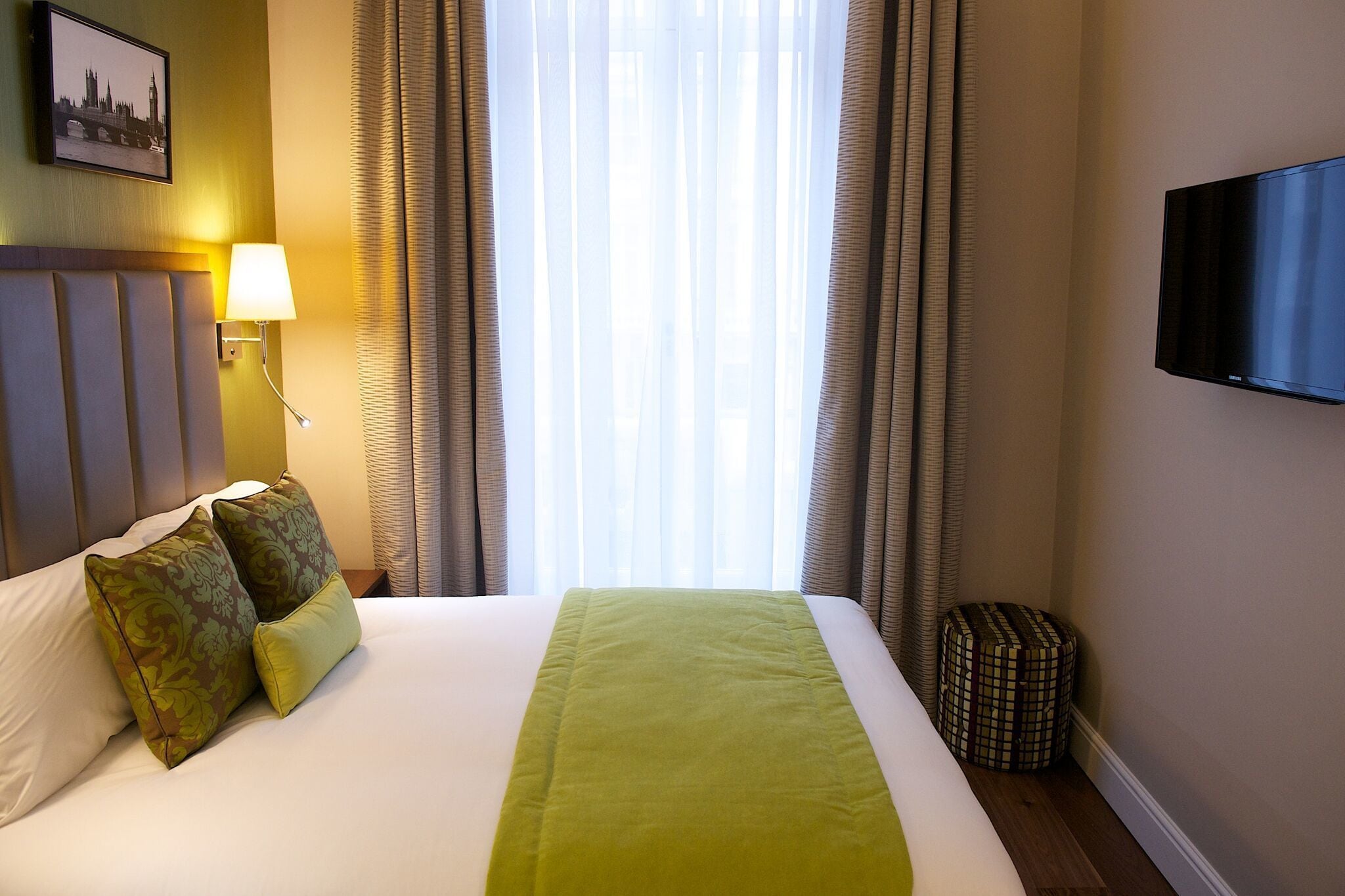 A hotel room with a bed, a television mounted on the wall, a lamp, and a window with curtains.