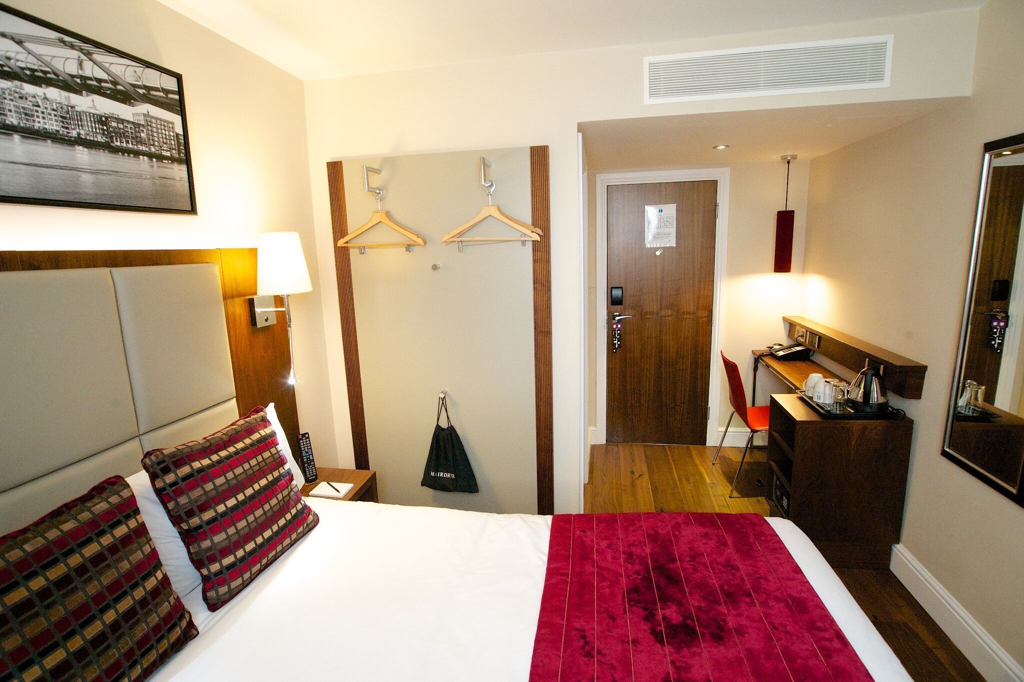 A hotel room with a bed, a desk, a chair, a hanging bag, and a door leading to another room.