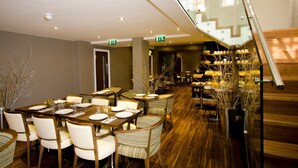A modern dining area with a long table set for a meal, surrounded by chairs, and a staircase leading to an upper level. - The Belgrave (London)