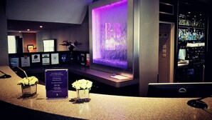 A hotel reception area with a large illuminated sign, a reception desk, framed awards, and a bar area. - The Belgrave (London)